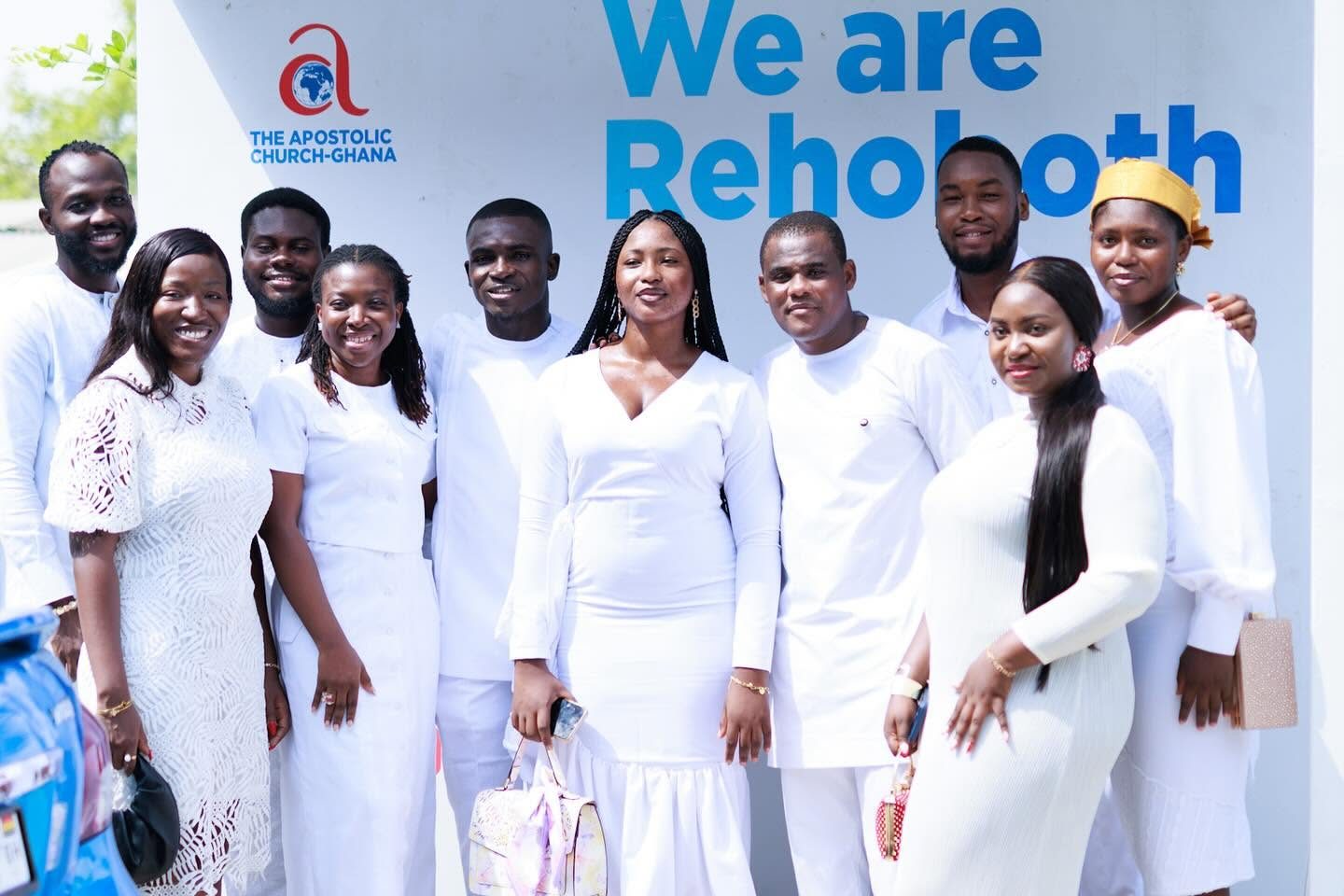 Members of the Rehoboth District dressed in white, standing together in front of the We are Rehoboth wall